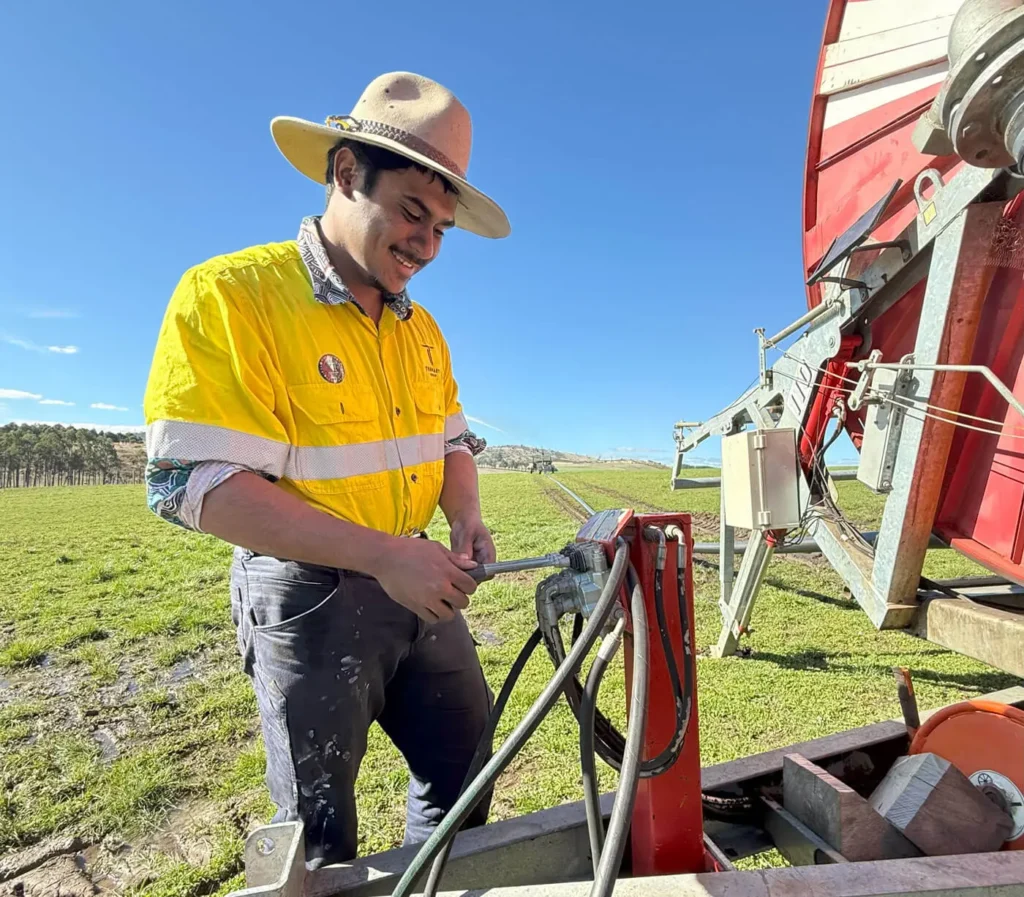 A young man in a bright yellow work shirt meticulously adjusts the machinery of a large center-pivot irrigation system in a vast, sun-drenched field